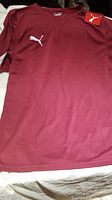 Front view of a burgundy Puma team shirt with white logo and tag on the left side of the collar.