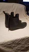 Side view of both new, grey Storm rain boots with elastic side panels and pull tabs, placed on beige surface.