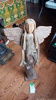 Full view of the wooden angel sculpture showing carved face, long arms, and driftwood wings on wooden base