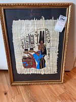 Framed papyrus artwork depicting an Egyptian figure with hieroglyphics inside a gold ornamental frame on hardwood floor.
