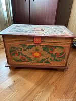 Front view of the wooden chest showing large floral carved and painted design with flower center and leafy scrolls in green, red, and yellow hues on a natural wood background.