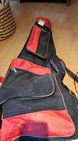 Red and black fencing bag with several compartments and zippers, lying on wooden floor.