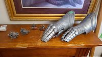 Wide shot of both articulated metal gauntlets next to four medieval-themed wall hooks on wooden tabletop