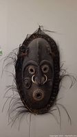 Full frontal view of the carved wood mask mounted on the wall, showing overall shape, bamboo frame, and raffia fringe.