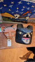 Two wall hanging masks displayed against a colorful painted backdrop. Left is the clay mask with large ears and visible teeth, right the black wooden mask with an open mouth and ears.