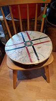 View of rain drum placed on wooden chair showing top painted design with intersecting colored lines and small perforations.
