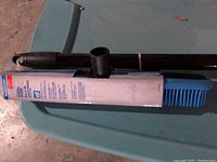 Photo showing a black telescopic broom handle with a blue Mastercraft rubber broom head attached, laying on a greenish surface.