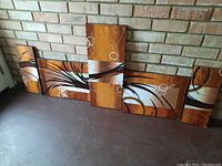 Set of five abstract decorative art panels arranged on a floor against a brick wall showing warm tones with curved black lines and white circular shapes.