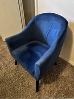 Blue velour accent chair against a beige wall and brown carpet flooring, showing upholstered seat, back, and armrests with dark wooden legs.