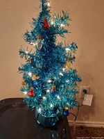 Blue small artificial Christmas tree with clear white and colored lights and small bulb ornaments on a stand