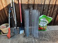 Photo showing garden tools including shovel, cultivator, hand brush, rolls of wire mesh, and bag of potting soil against wooden fence.