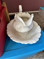 White ceramic pitcher with intricate embossed floral and scroll designs placed inside a large scalloped edge white ceramic bowl.