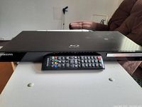 Front view of one Samsung black Blu-ray DVD player with remote placed on top on white surface.