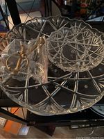 2-tier clear crystal serving platter with metal handle and accompanying crystal pieces, displayed on shelf surface.