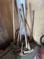 Overall view of multiple axes, picks, mauls and handles leaning against wall showing condition and quantity.