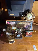Overall view of assorted mantle clocks, a decorative wall clock, and a GE clock radio arranged on a floor.