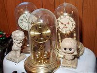 Three anniversary clocks each with a glass dome, plus two ceramic figurines with sentimental inscriptions. Clocks show different dial designs, one gold-tone, others white with floral or numeric faces.
