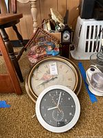 Overview of several German style miniature grandfather clocks, Helio weather station clock, bird sound clock, decorative clock face, and miscellaneous items like string and electrical tape on floor near chair.