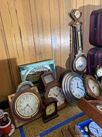 Photo showing multiple clocks including a large wooden wall barometer and thermometer, round wall clocks, square mantel clocks, small brass dial clocks, and boxed indoor/outdoor thermometer