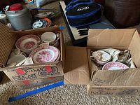 Boxed ironstone dinnerware pieces with red transferware design including plates, cups, and bowls packed in newspaper inside two cardboard boxes.