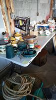 Photo of folding table with various kitchenware and miscellaneous items placed on top including rope, containers, and tape. Background is a garage setting with cinder block walls.