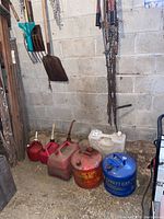 Photo of seven metal gas cans lined up on the floor including red, white, and blue cans, all showing rust and heavy use, hanging rusted chains on the wall behind.