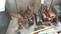 Photo showing multiple hydraulic jacks, jack stands, and handles grouped on a concrete floor against a cinder block wall. Items show surface rust and wear.