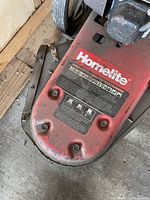 Close-up photo of the front deck of the lawnmower showing the red metal surface with Homelite branding and warning labels. Shows wear and some dirt.