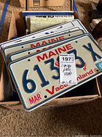 Box containing multiple Maine license plates with Vacationland and lobster design dated early 1990s, stacked in box with folded road maps underneath