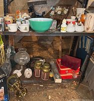 Overview of shelf with turquoise Pyrex bowl, various decorative cups, piggy banks, condiment server, salt and pepper shakers, and vintage brass outdoor light fixture on floor.