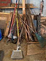 Photo showing a variety of long-handled gardening tools including shovels, rakes, hoes, and wooden handles stacked against a wall in a garage.