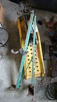 Pair of yellow metal automotive ramps with holes in surface and green metal stand and jack in garage setting. Visible rust and weathering.