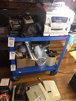 Blue metal rolling cart with two shelves holding various items including electronics and boxes. Cart frame is bright blue metal with caster wheels.