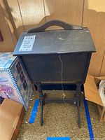 Dark wooden sewing box on a stand with a hinged lid and handle, showing some dirt and stains, placed against a wood paneled wall with boxes nearby.