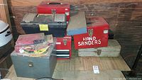 Photo showing a collection of multiple toolboxes in various colors including red, gray, and blue, some labeled 'HAND SANDERS.'