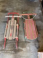 Top view of two vintage sleds placed on a wooden floor. One is a Flexible Flyer with natural wood slats and red metal frame. The other is a wooden sled with red runners and a green handlebar.