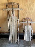 Full view of both vintage sleds leaning against a wooden wall, showing their wooden slats and rusted metallic frames.