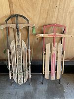 Photo showing two vintage wooden sleds, one Speedway branded and one Fasttrack branded, positioned upright against a wooden wall.