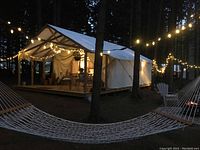 Canvas glamping tent in forest at dusk with string lights, hammock in foreground and Adirondack chair