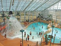 Indoor waterpark view showing wave pool, tipping bucket feature, lazy river and surrounding seating