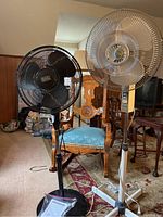 Full view of two standing oscillating fans, one black Black+Decker on the left and one white Windmere fan on the right with damaged front grille, in a room with wood paneling and furniture.