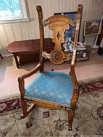 Full view of the carved wooden rocking chair showing front profile, blue fabric seat, and wood grain.