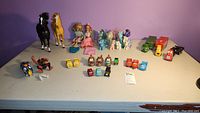 Overview of lot showing all items on table including horses, My Little Pony figures, Barbie dolls, toy trucks, and small cars.
