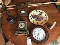 Photo showing four items on a wooden table: a metal lamp base, a silver ornate mantel clock, a wooden box mantel clock, and a round rooster-themed wall clock.