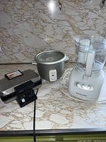 Photo showing three kitchen appliances on a marble countertop: Cuisinart pizzelle maker on the left, Farberware small crockpot in the middle, KitchenAid food processor on the right.