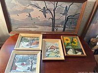 Five framed paintings displayed on a table and leaning against a wall. Includes large painting of birds and bare trees, winter landscapes with barns, and sunflowers.