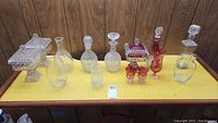 Full view of nine assorted glass items including three covered candy dishes, five decanters with stoppers, and one vase, all placed on a yellow surface showing need for cleaning.