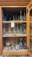 Three shelves of glasses inside a wooden cabinet showing the green textured glasses inverted on the top shelf, clear stemware on the middle shelf, and various clear glasses including decanter on the bottom shelf.