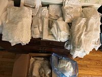 Photo showing multiple folded lace and crochet curtain panels and valances in off-white color stacked on wooden furniture, along with a box containing additional fabric pieces.