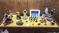 Full lot overview showing various lighthouse figurines, snow globe, framed pictures on yellow table and wood panel background.